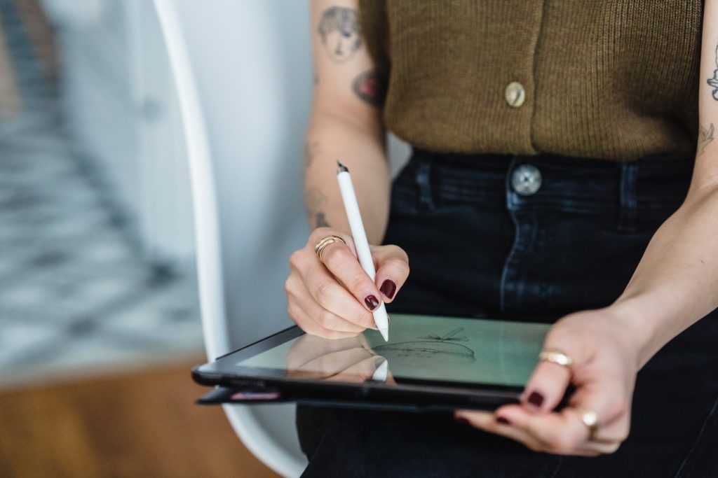 close up image of woman's torso as she is holding an ipad and stylus. Woman is tattood. She is writing notes on the ipad with her stylus.