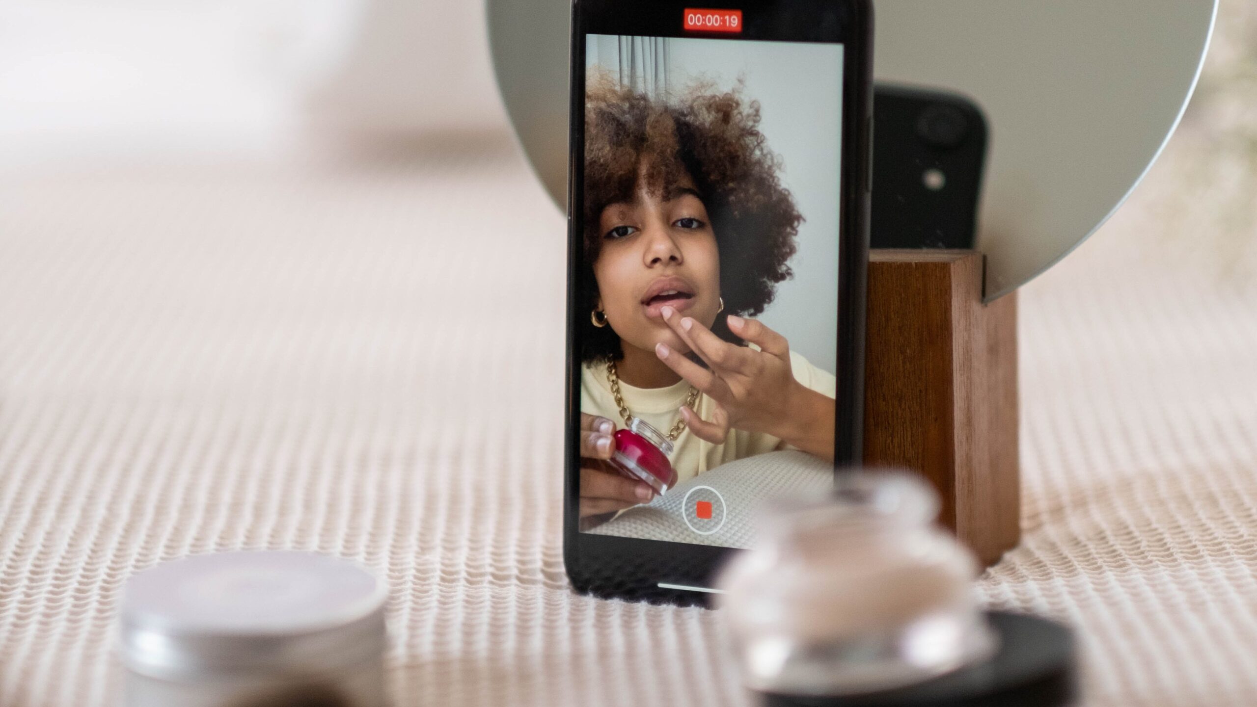 Peering at a phone propped up against a mirror as a black woman with kinky hair puts on lipgloss while recording herself on the phone.